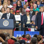 Two politicians speaking at a political rally with a large crowd in attendance