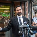 NYC Mayor’s Free Childcare Scam Exposed Man in a suit speaking at a press conference with microphones in front of him