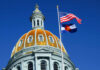 The dome of a state capitol building with flags flying against a blue sky