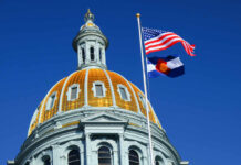 The dome of a state capitol building with flags flying against a blue sky