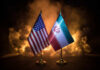 Flags of the United States and Iran displayed on stands with a smoky background