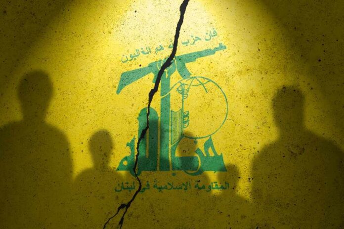 shutterstock_2373422413 (1).jpg Silhouettes of individuals against a yellow wall featuring Hezbollah's emblem