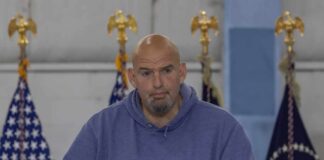Man in a blue hoodie speaking at a podium with U.S. flags behind