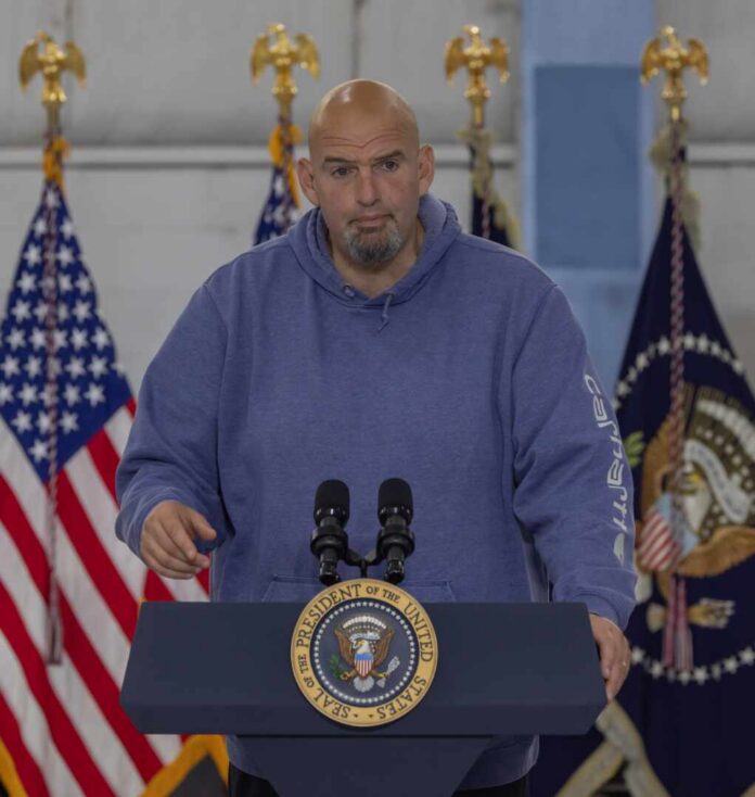 Man in a blue hoodie speaking at a podium with U.S. flags behind