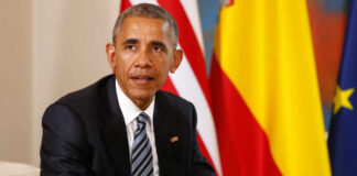 Man in a dark suit seated with flags in the background