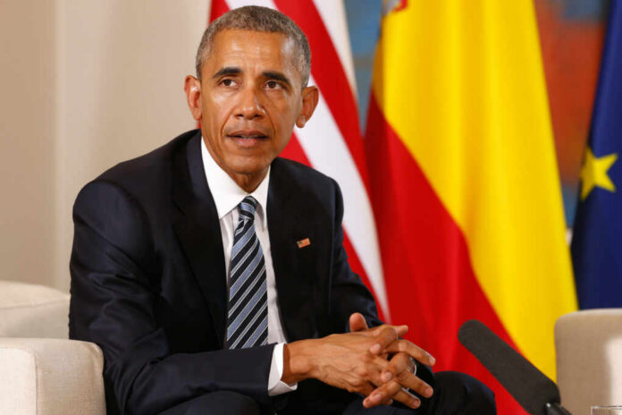 Man in a dark suit seated with flags in the background
