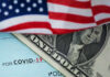 A dollar bill partially covered by an American flag and a COVID-19 stimulus check