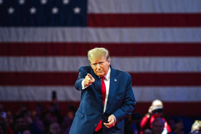 trumpid2455396913jpg Man points in front of American flag backdrop.