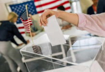 Hand placing ballot in box with American flag nearby