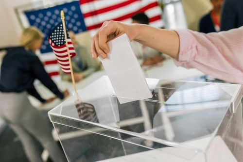 Hand placing ballot in box with American flag nearby