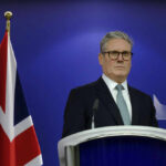 Man in suit at a podium, British flag behind.
