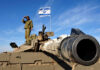 Who’s Really Running This Middle East War? Soldier saluting from atop a tank with an Israeli flag in the background
