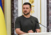 Man speaking at podium with microphones, Ukrainian flag behind.