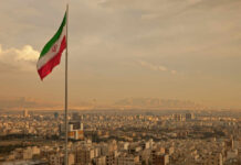 Five-Hour U.S. Blitz Hits Iran Iranian flag waving over a city skyline with mountains in the background