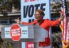 Speaker at outdoor rally behind podium with vote signs