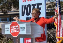 Speaker at outdoor rally behind podium with vote signs