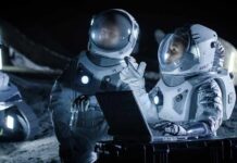 Two astronauts in spacesuits review data on a laptop on a moonlike surface at night