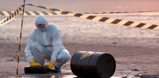 Hazmat worker kneeling near a fallen barrel at a cordoned-off site