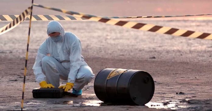 Hazmat worker kneeling near a fallen barrel at a cordoned-off site