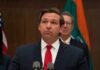Ron DeSantis at a press conference podium in a suit and red tie