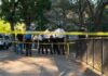 Police officers gather behind yellow tape on a tree-lined sidewalk