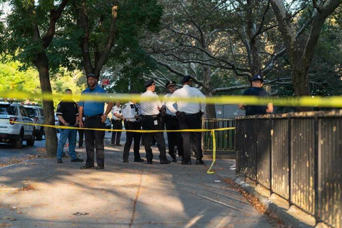 Police officers gather behind yellow tape on a tree-lined sidewalk